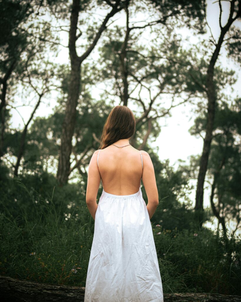 Home A woman in an elegant backless white dress stands in a tranquil forest setting.